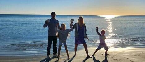 Une famille de quatre personnes se trouve sur une plage, le soleil étant bas à l’horizon et projetant de longues ombres.