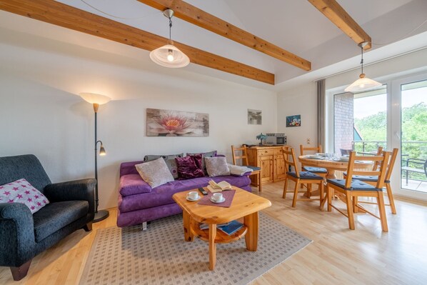 Un salon chaleureux avec un canapé, une table basse et une table à manger avec des chaises. On y trouve une cheminée, une fenêtre offrant une belle vue et un plafond avec des poutres apparentes.