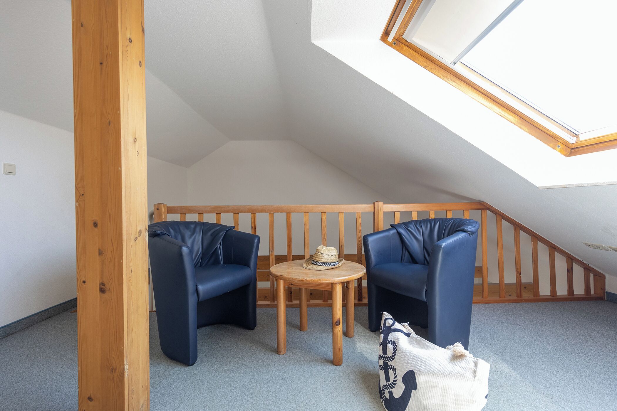 A small, cozy room with a sloped ceiling, wooden beams, two blue chairs, a small round table with a hat, and a patterned cushion.