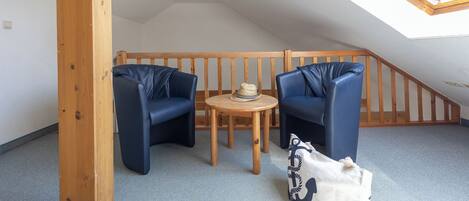 A small, cozy room with a sloped ceiling, wooden beams, two blue chairs, a small round table with a hat, and a patterned cushion.
