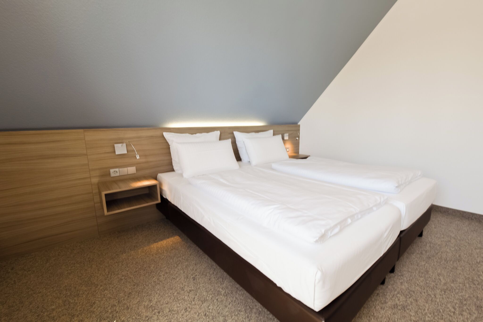 Moderni hotellihuone, jossa on puinen sängynpääty, kaksi sänkyä, yöpöytä ja seinään kiinnitetty valaisin.