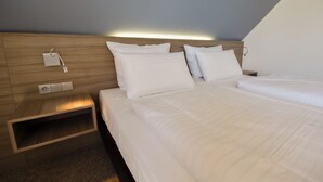 A modern hotel room with a wooden headboard, two beds with white linens, a bedside table, and a wall-mounted light. - Double room 112. Modern design and close to the beach. (Wurster Nordseeküste)