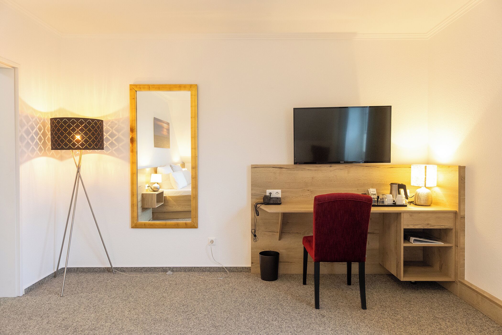 Ein Hotelzimmer mit Schreibtisch, Fernseher, rotem Sessel, Stehlampe und Spiegel.