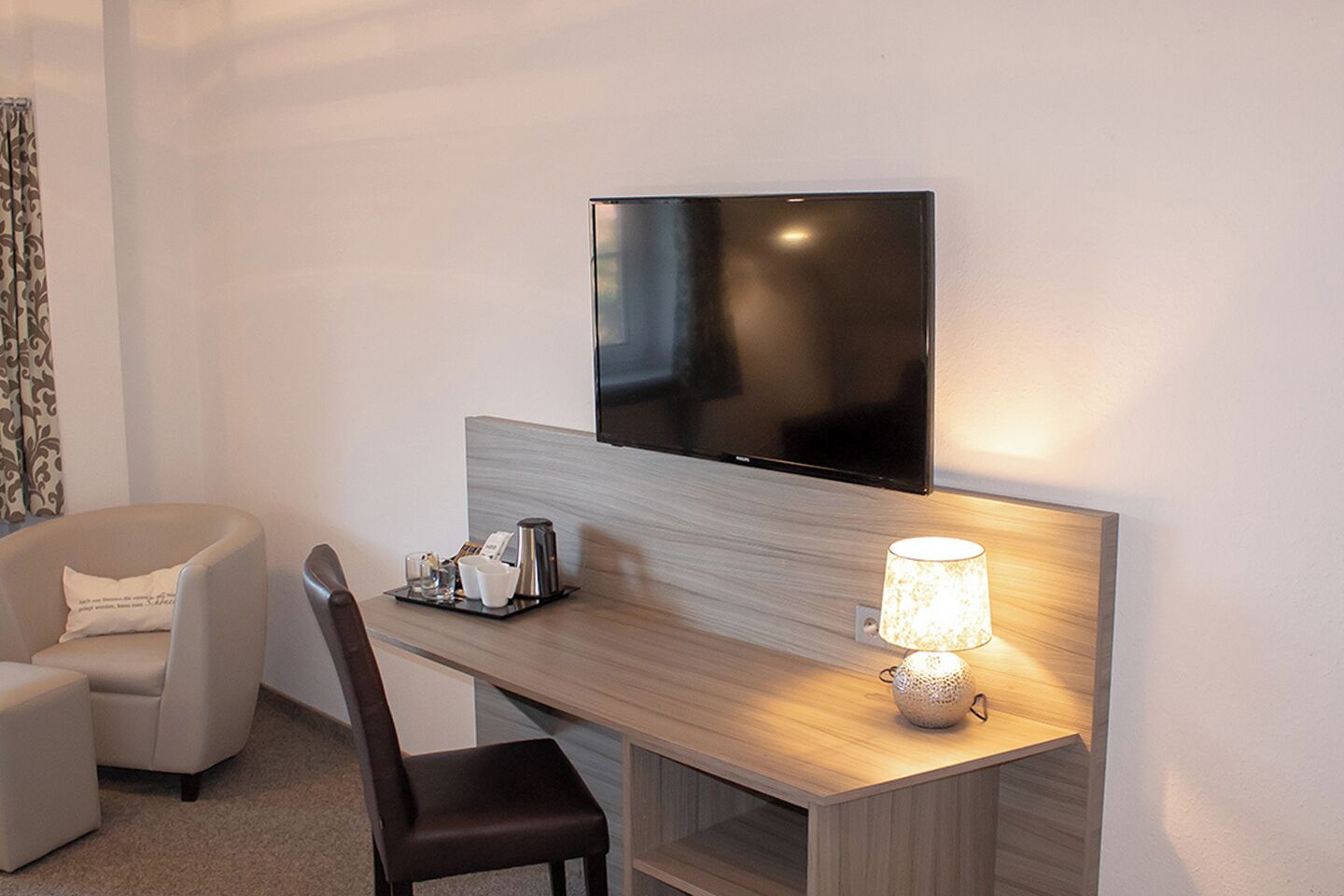 Ett hotellrum med en platt-TV som är monterad på väggen, ett skrivbord i trä med en lampa, en stol och ett litet bord med en kaffekanna och koppar.