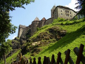 A castle-like building with multiple towers, situated on a grassy hill with a wooden fence in the foreground.
