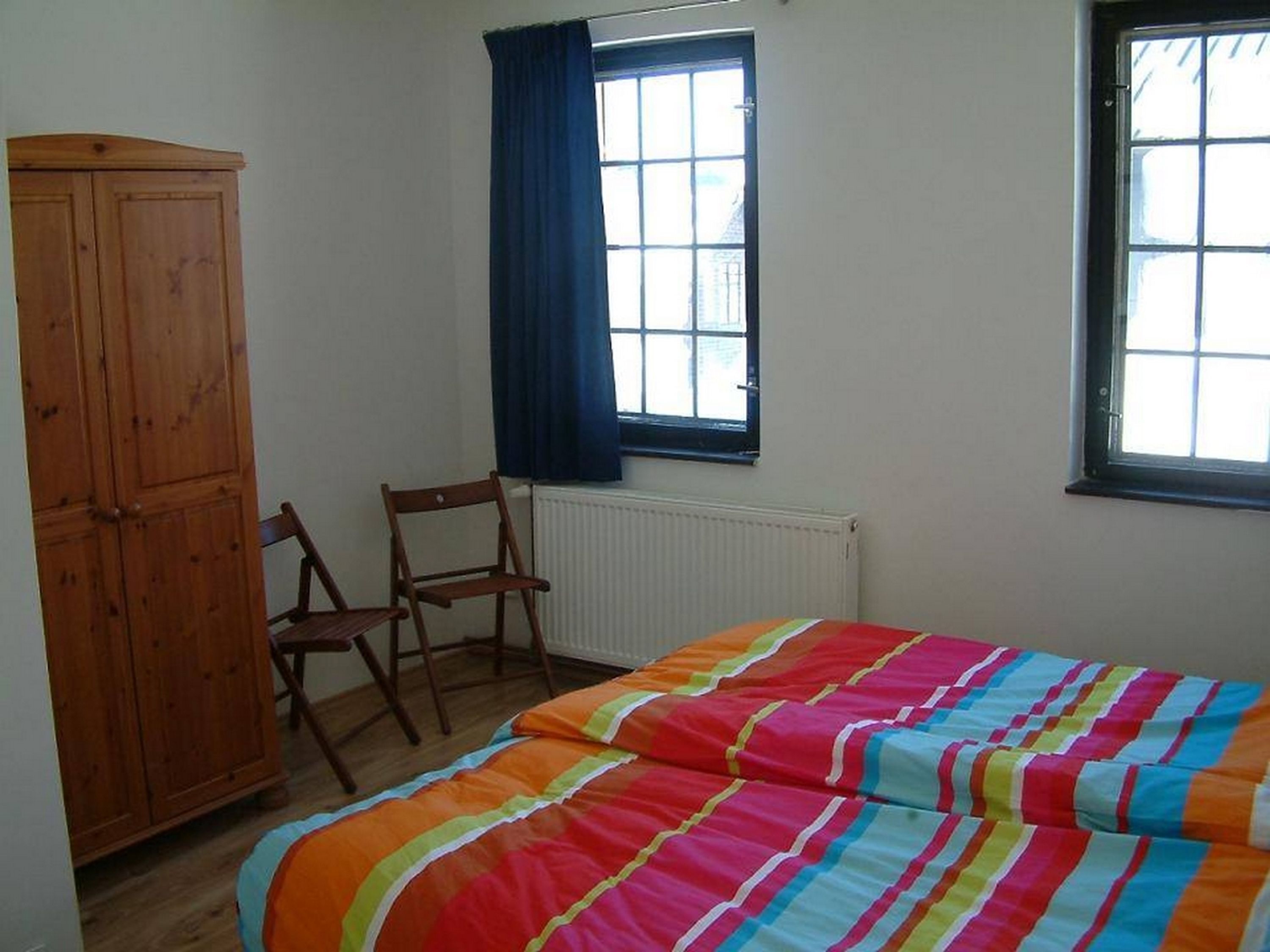 A bedroom with a bed, a wooden wardrobe, two chairs, and two windows.