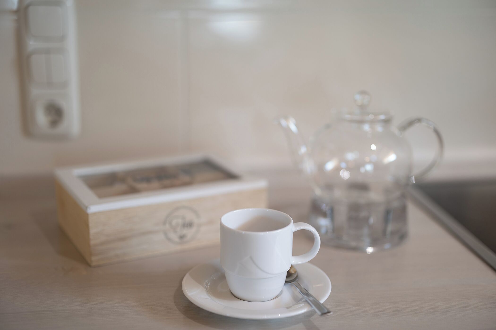 A white cup on a saucer with a spoon, a glass teapot, and a wooden box with a picture on a table.