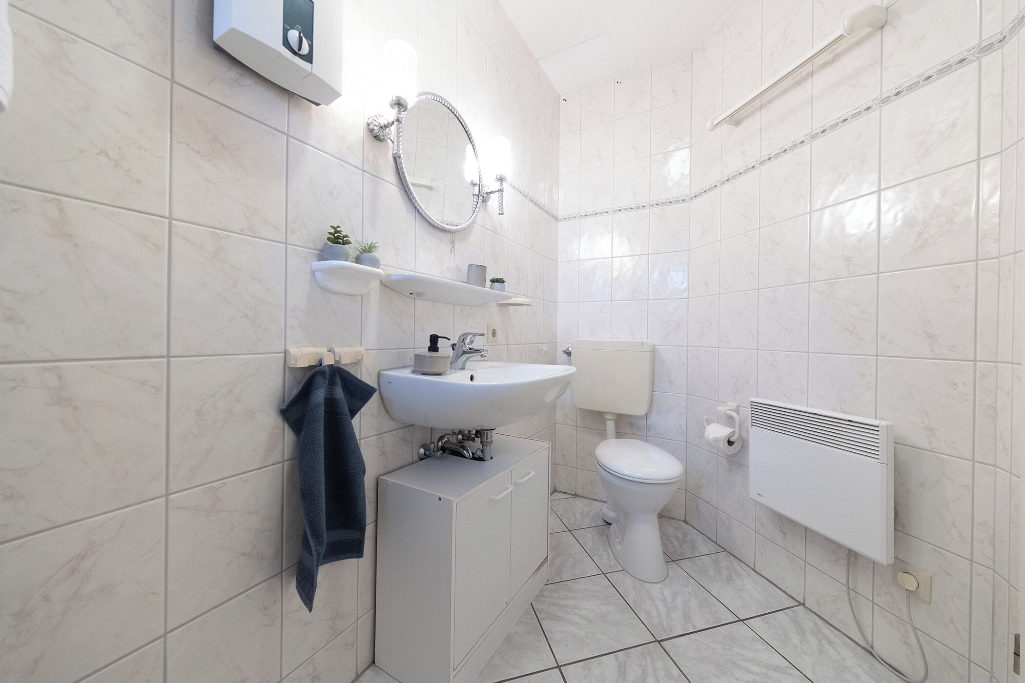 A bathroom with a white sink, toilet, and radiator. There are shelves above the sink and a towel hanging on the wall.