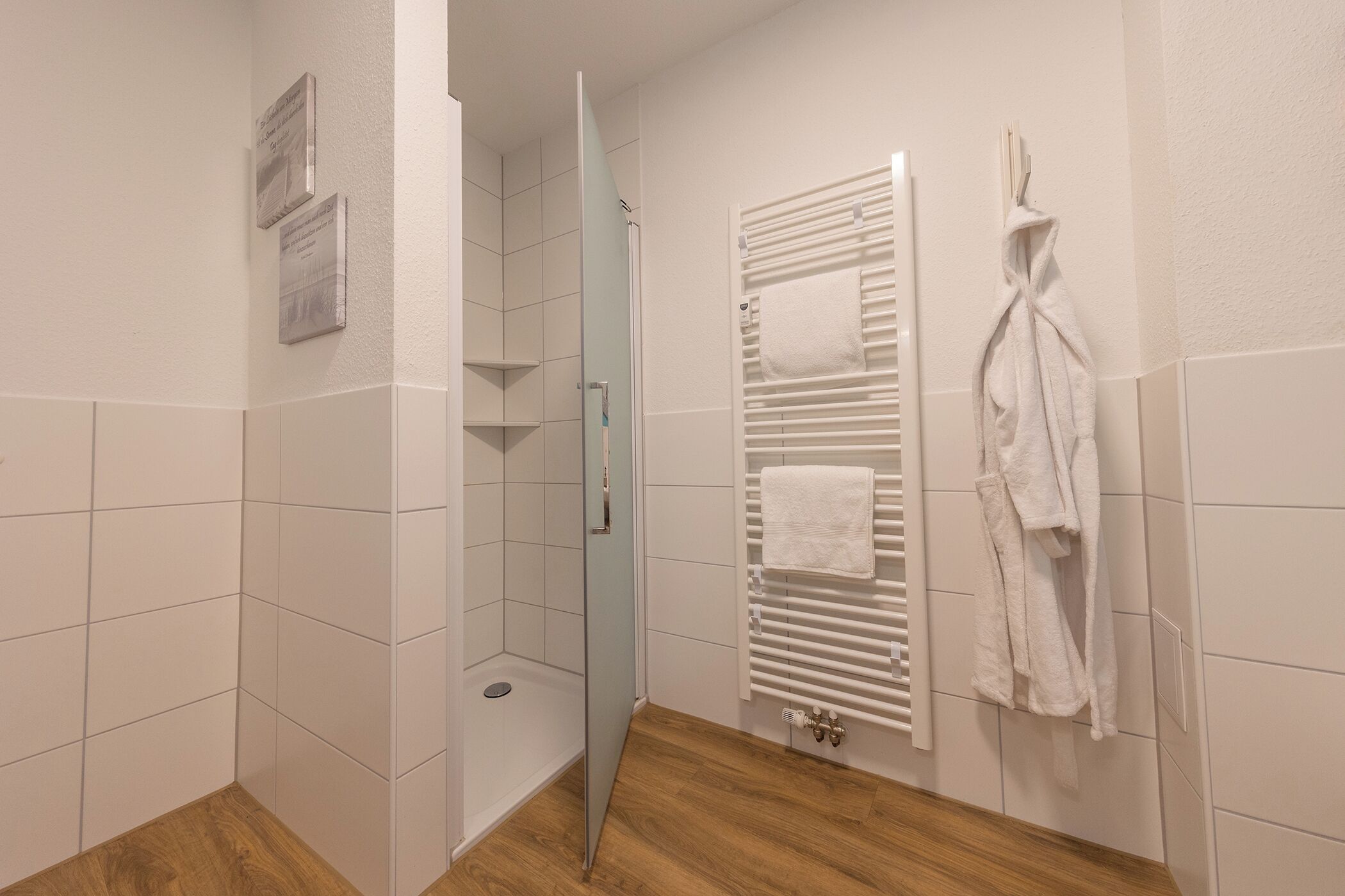 A bathroom with white tiles, a glass shower enclosure, a towel rack, and a wooden floor.