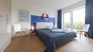 Une chambre moderne avec un grand lit, deux chaises blanches, une table de chevet, un dispositif de sauvetage fixé au mur et une grande fenêtre avec des rideaux.