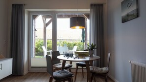 A dining area with a round table, four chairs, a hanging light fixture, and a view through large windows.