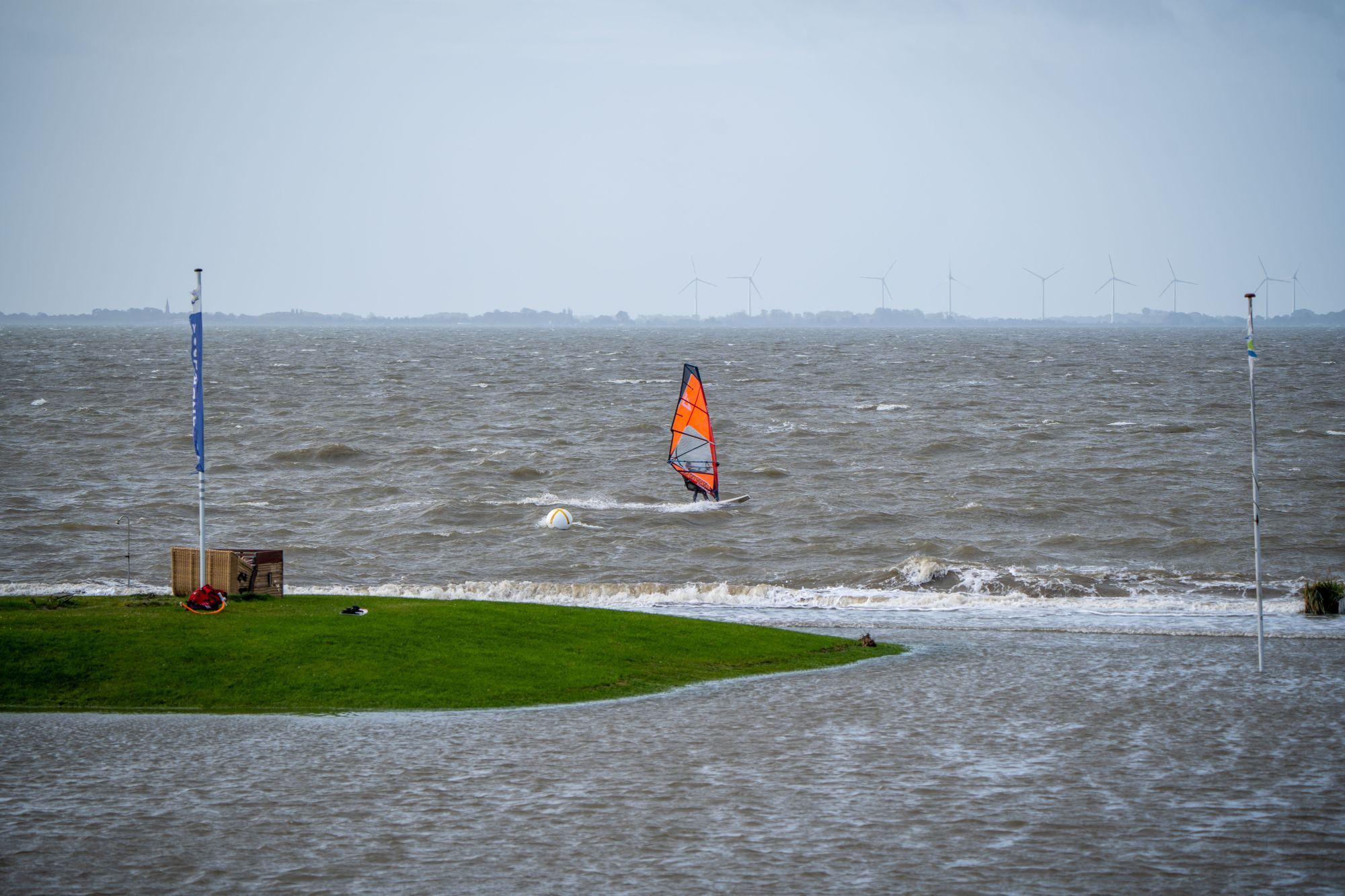 Een windsurfer op het water, met een grasveld en een vlaggenmast op de voorgrond.