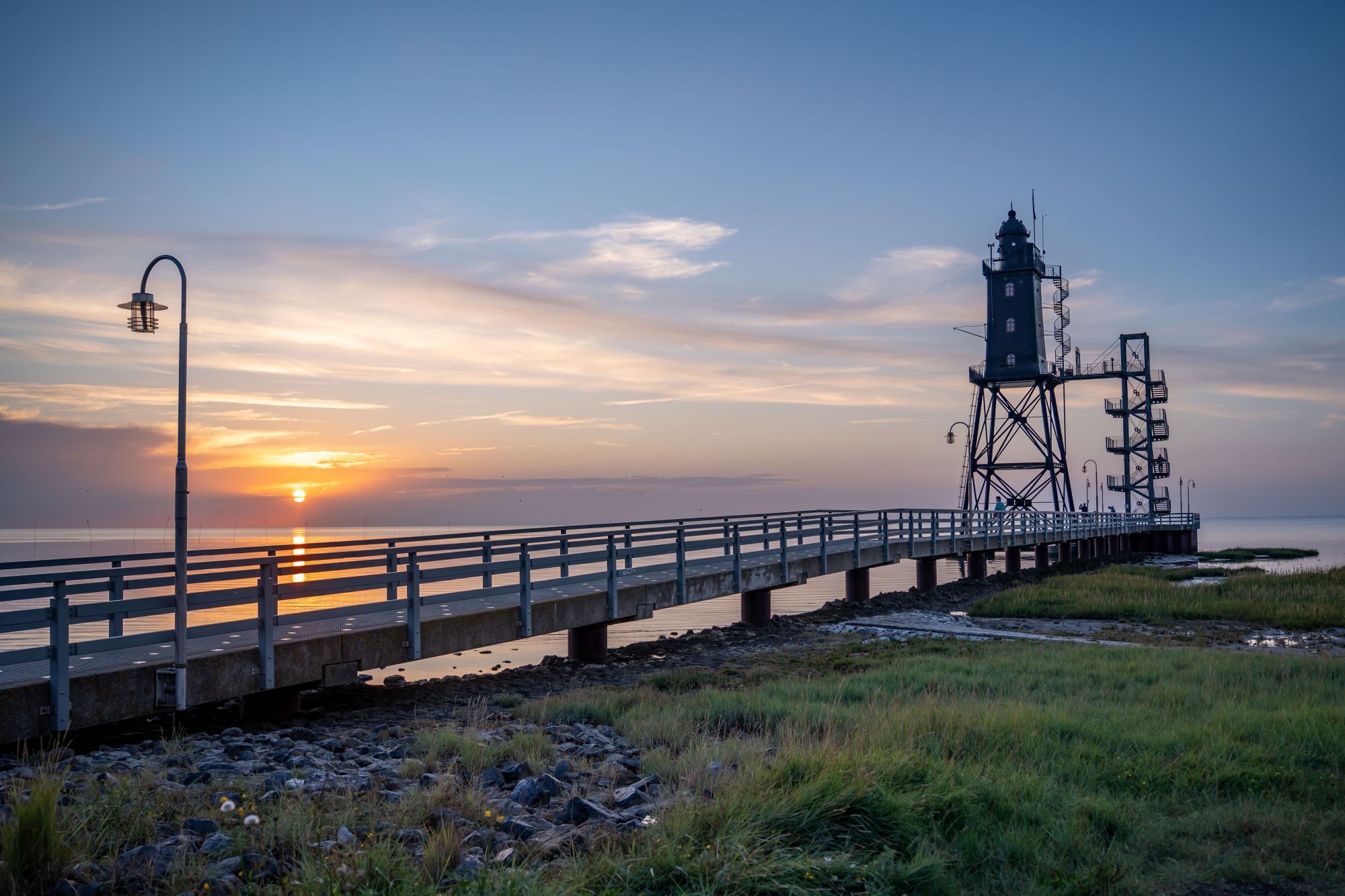 Eine Steg, der sich über ein Gewässer erstreckt, mit einem Leuchtturm im Hintergrund.