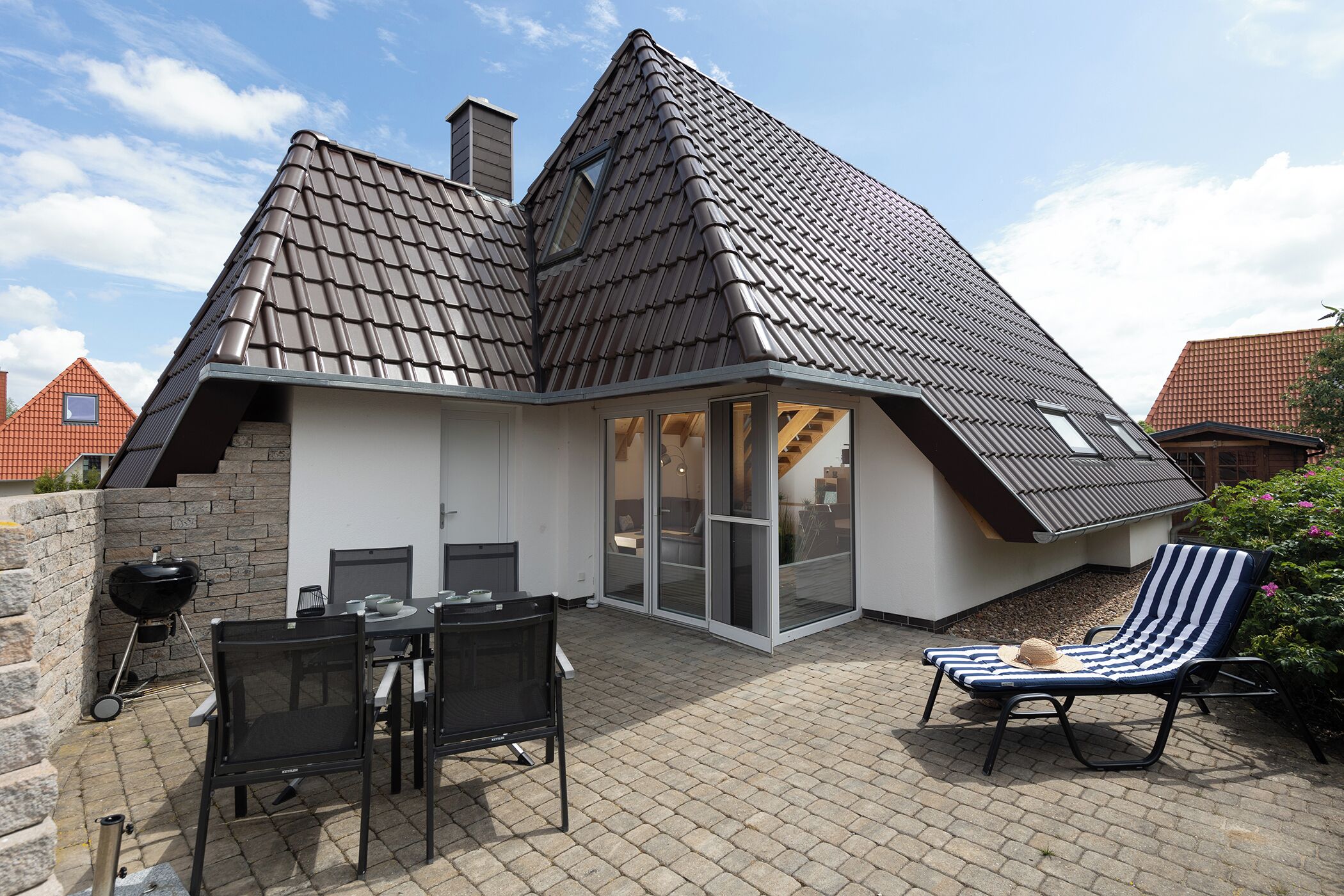 A house with a sloped roof, a patio with a table and chairs, and a lounge chair.