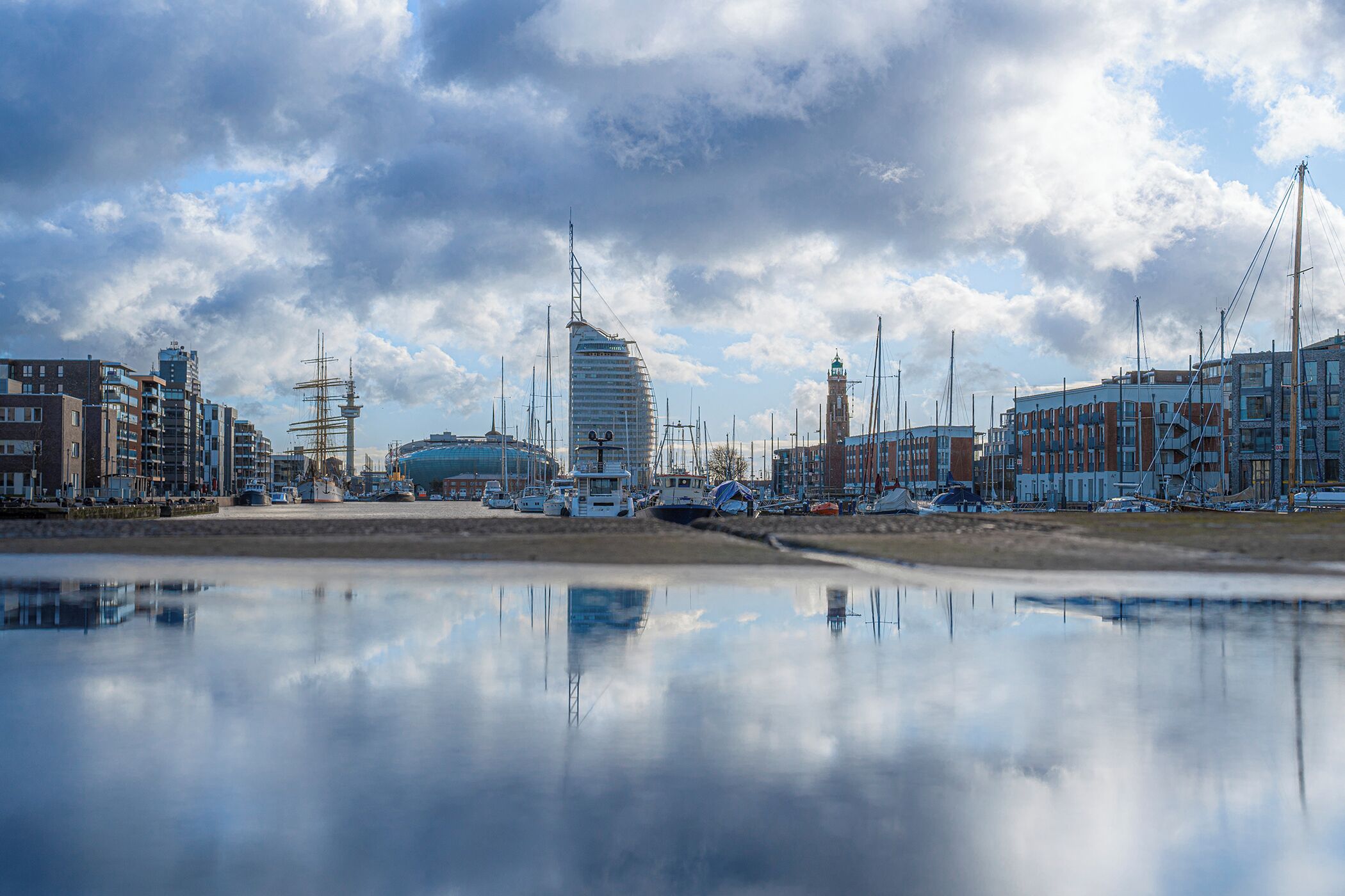 Eine Uferstadt mit moderner Architektur, darunter ein markanter, geschwungener Wolkenkratzer, dessen Spiegelbild sich in ruhigem Wasser zeigt.