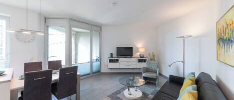 Un salón moderno con un sofá de cuero negro, una mesa de centro de vidrio y un comedor con una mesa blanca y sillas oscuras.