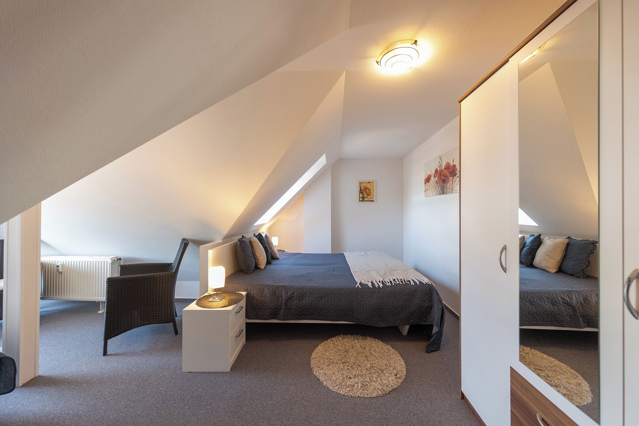 A bedroom with a sloped ceiling, a bed with a gray blanket, a chair, a small table, and a radiator.