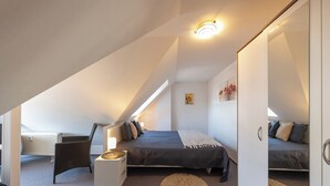 Ein Schlafzimmer mit schrägen Wänden, einem Bett mit grauer Decke, einem Sessel, einem kleinen Tisch und einer Heizung.