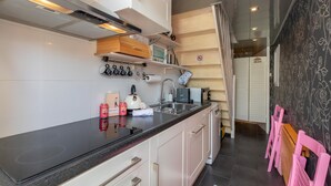 A modern kitchen with a black countertop, stainless steel sink, and white cabinets. There are shelves with kitchen items and a wooden cutting board. - Studio De Zandhut - Swaalingestraat 78 Zoutelande (Zoutelande)