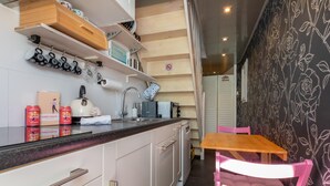 A compact kitchen with white cabinetry, a wooden countertop, and a small dining area with a pink chair. - Studio De Zandhut - Swaalingestraat 78 Zoutelande (Zoutelande)