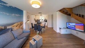 A modern living room with a sofa, coffee table, and a TV displaying 'Brokstedt'. A staircase and kitchen are visible in the background. - Vacation terraced house Juist. Comfortable and inviting. (Wurster Nordseeküste)