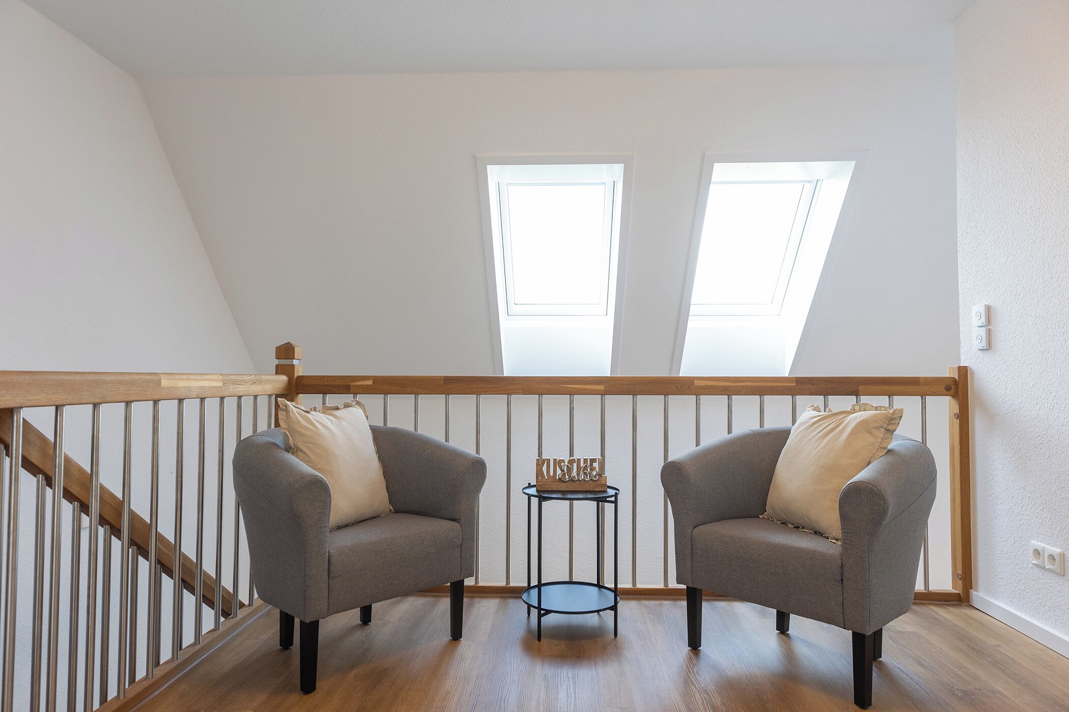 A loft bedroom with two grey armchairs, a small round table, and a wooden railing.