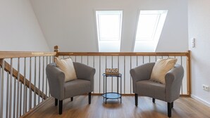 A loft bedroom with two grey armchairs, a small round table, and a wooden railing.