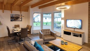 A modern living room with a dining area, wooden ceiling beams, large windows, a television, and a wooden coffee table.