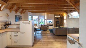 A spacious living area with wooden beams, a kitchen with white cabinets, a dining area, and a large window offering a view of the outdoors.
