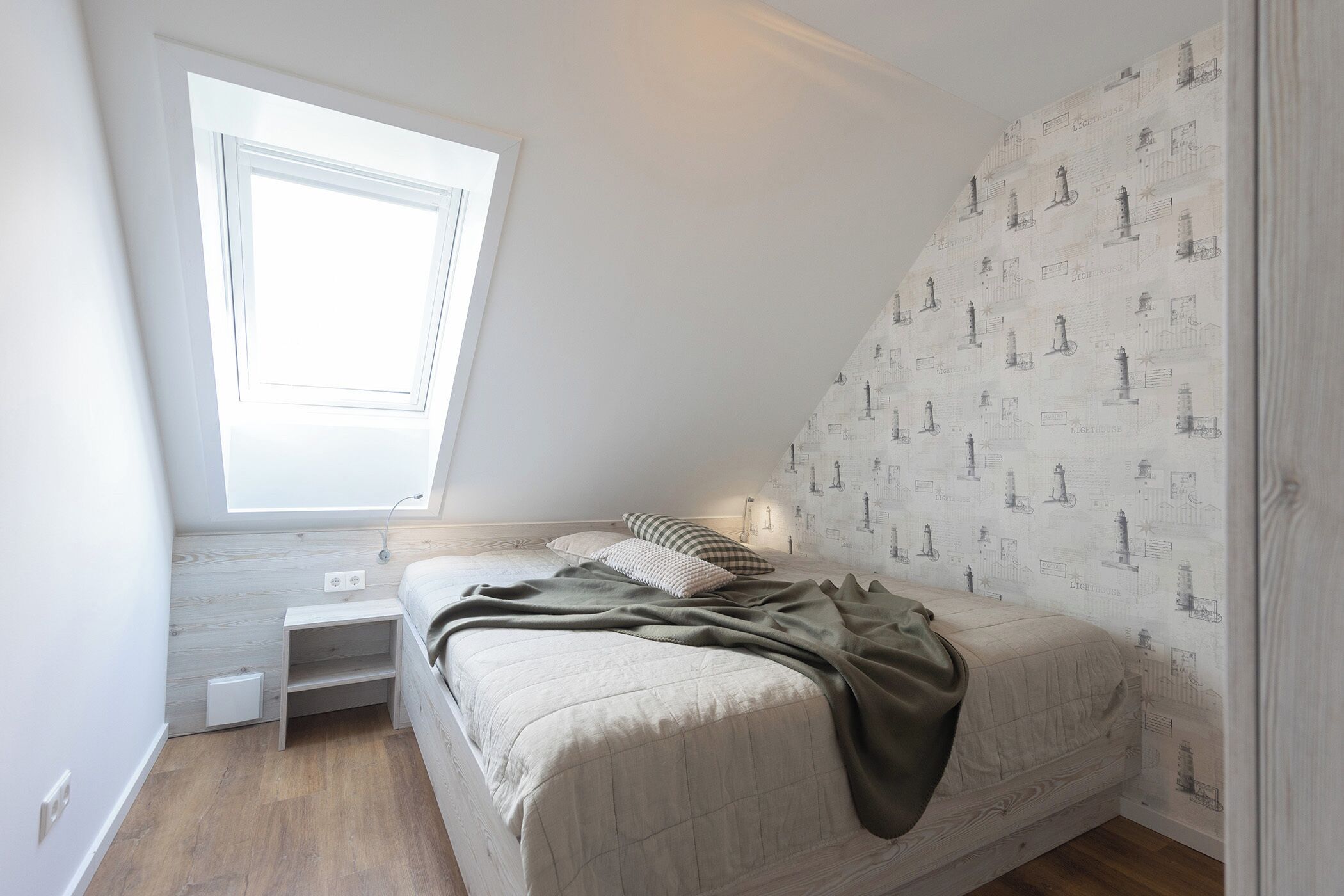 A bedroom with a sloped ceiling, a window, a bed with a blanket, and a wall with a lighthouse pattern.