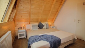 Un dormitorio acogedor con techo inclinado, paredes de madera, una cama con almohadas, una mesita de noche con una lámpara y un radiador.