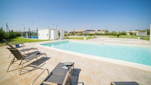 Piscina com espreguiçadeiras e uma pequena mesa, cercada por área ajardinada sob céu claro.