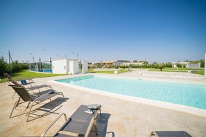 Una piscina con sdraio e un tavolino, immersa in un’area verde curata, sotto un cielo sereno.