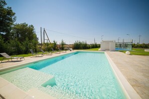 Una piscina rettangolare con acqua cristallina, circondata da un solarium piastrellato. Si vedono lettini e una piccola struttura nelle vicinanze.