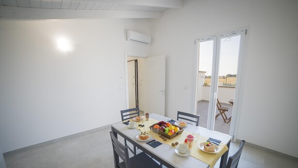 Uma área de refeições com uma mesa posta para quatro pessoas, incluindo pratos de comida, copos e uma cesta de frutas.