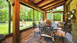 A covered patio with a wooden ceiling, a table with chairs, and a view of a garden.