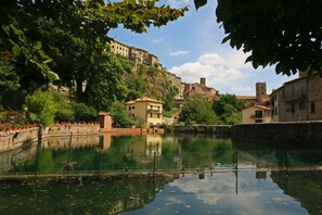 A calm river with buildings on a hillside, a clear sky, and trees in the foreground.