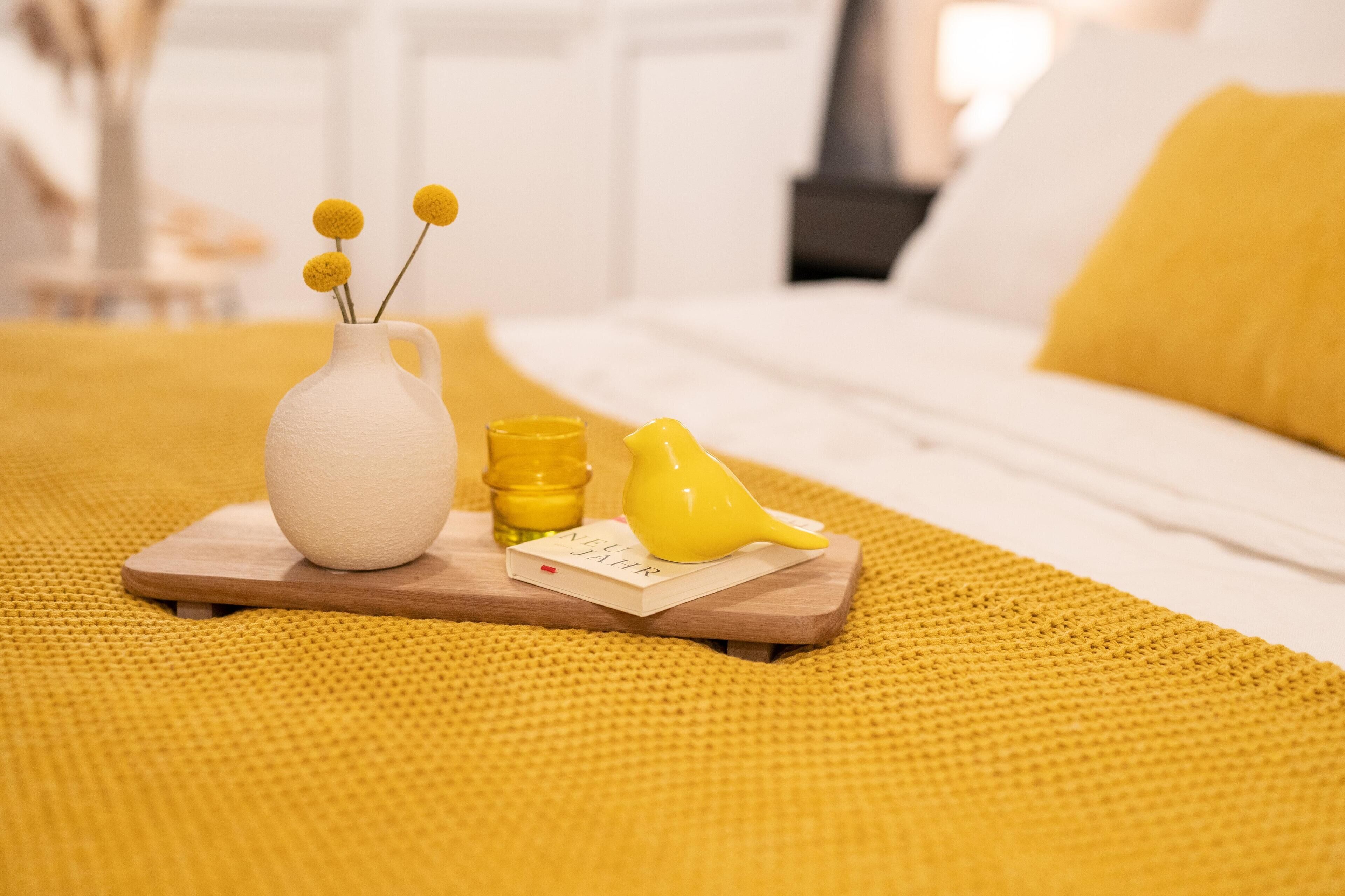 A bed with a yellow pillow, a white vase with yellow flowers, a yellow bird figurine, and a wooden tray with a small book and a yellow bottle.