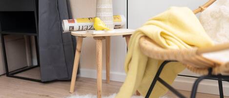 Una habitación acogedora con una silla de mimbre, una pequeña mesa de madera con una revista y un objeto amarillo, y un par de pantuflas sobre una alfombra mullida.