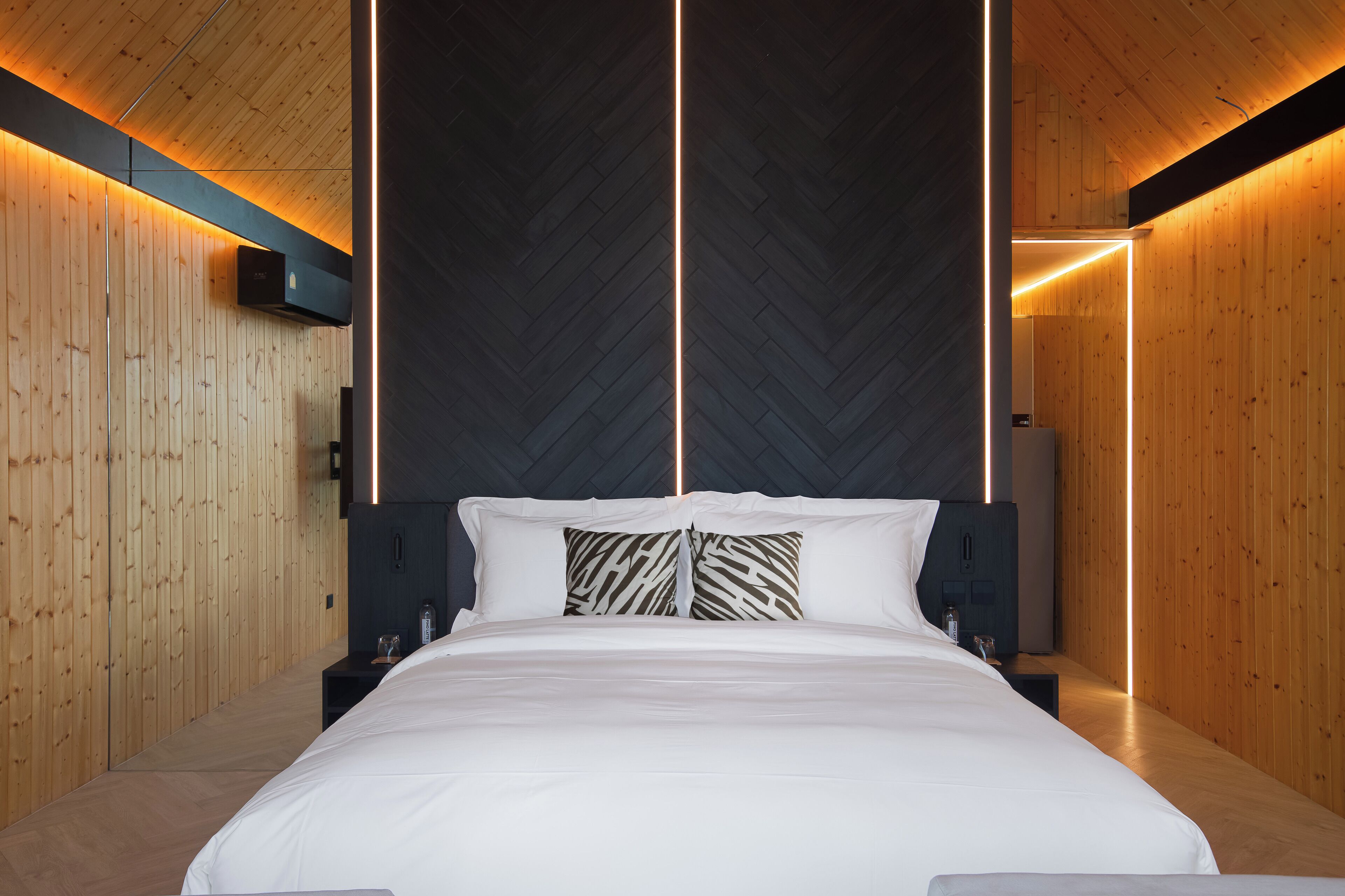 A modern hotel room with a large bed, wooden walls, and a central light feature.
