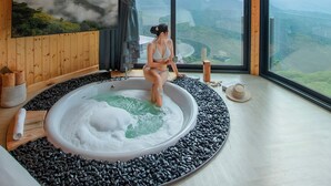 Une salle de spa dotée d’une grande baignoire entourée de galets noirs, d’un mur en bois et d’une grande fenêtre offrant une vue sur les montagnes.