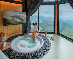 A spa room with a large bathtub surrounded by black pebbles, a wooden wall, and a large window offering a view of mountains.