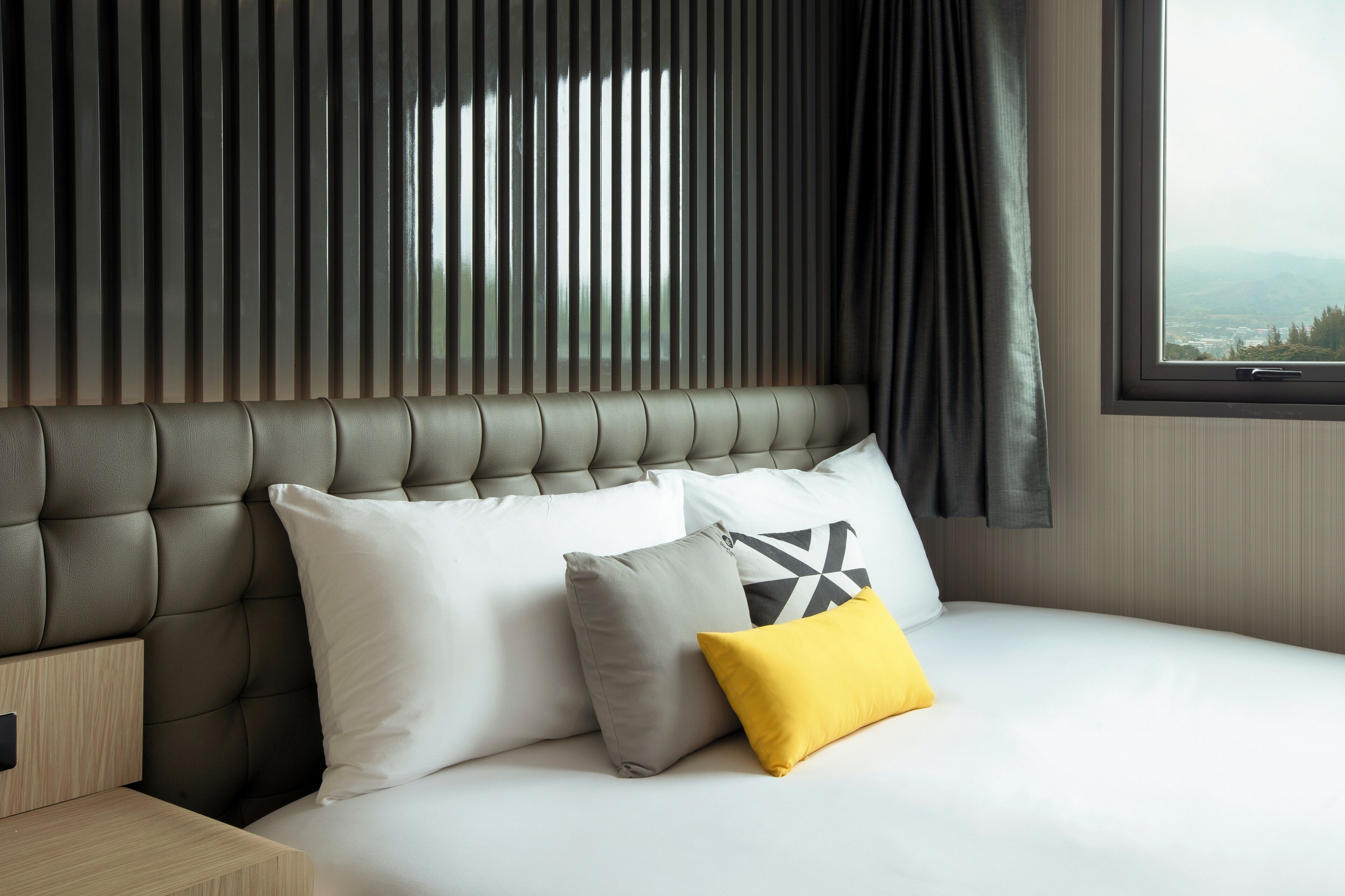 A modern hotel room with a tufted headboard, a large window with a view, and a neatly made bed with pillows.