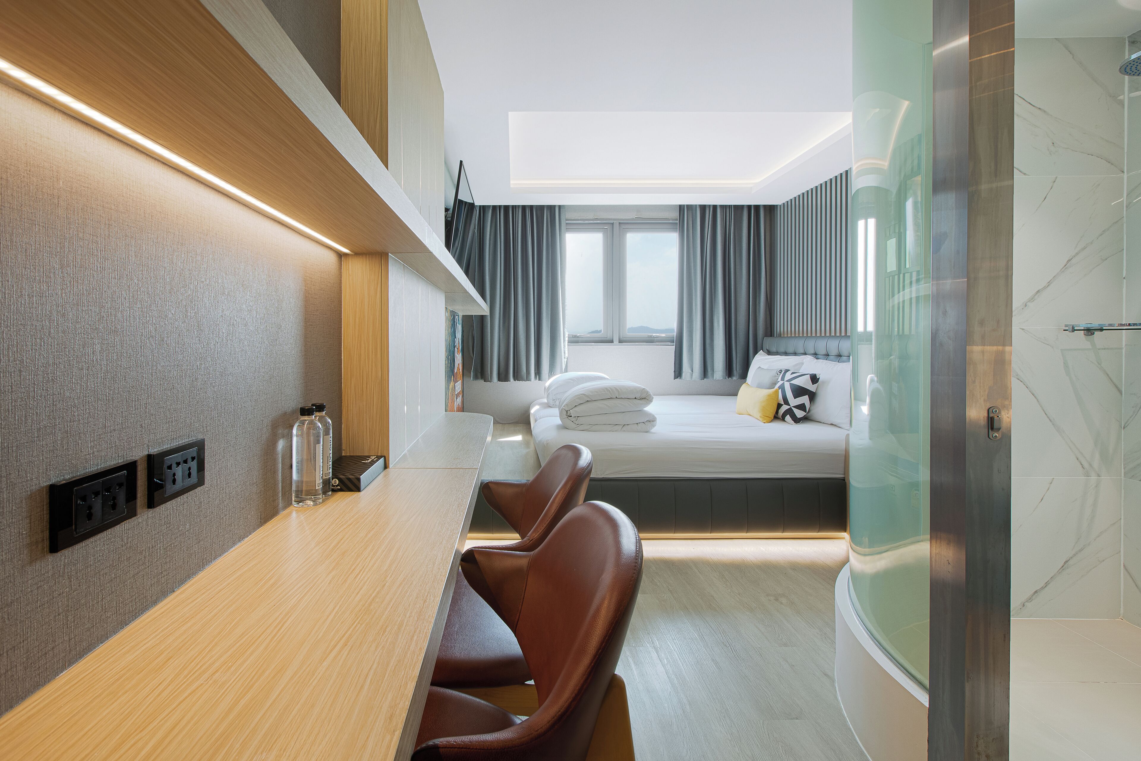 A modern hotel room with a wooden desk, leather chairs, a bed with a view, and a glass shower enclosure.