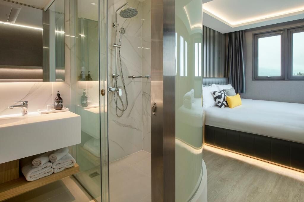 Un baño moderno con ducha envuelta en vidrio, tocador blanco con encimera de mármol y una cama con cabecero y almohadas.