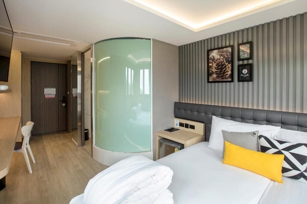 A modern hotel room with a curved glass partition, a bed with white and yellow pillows, a small bedside table, and a headboard with a tufted design.