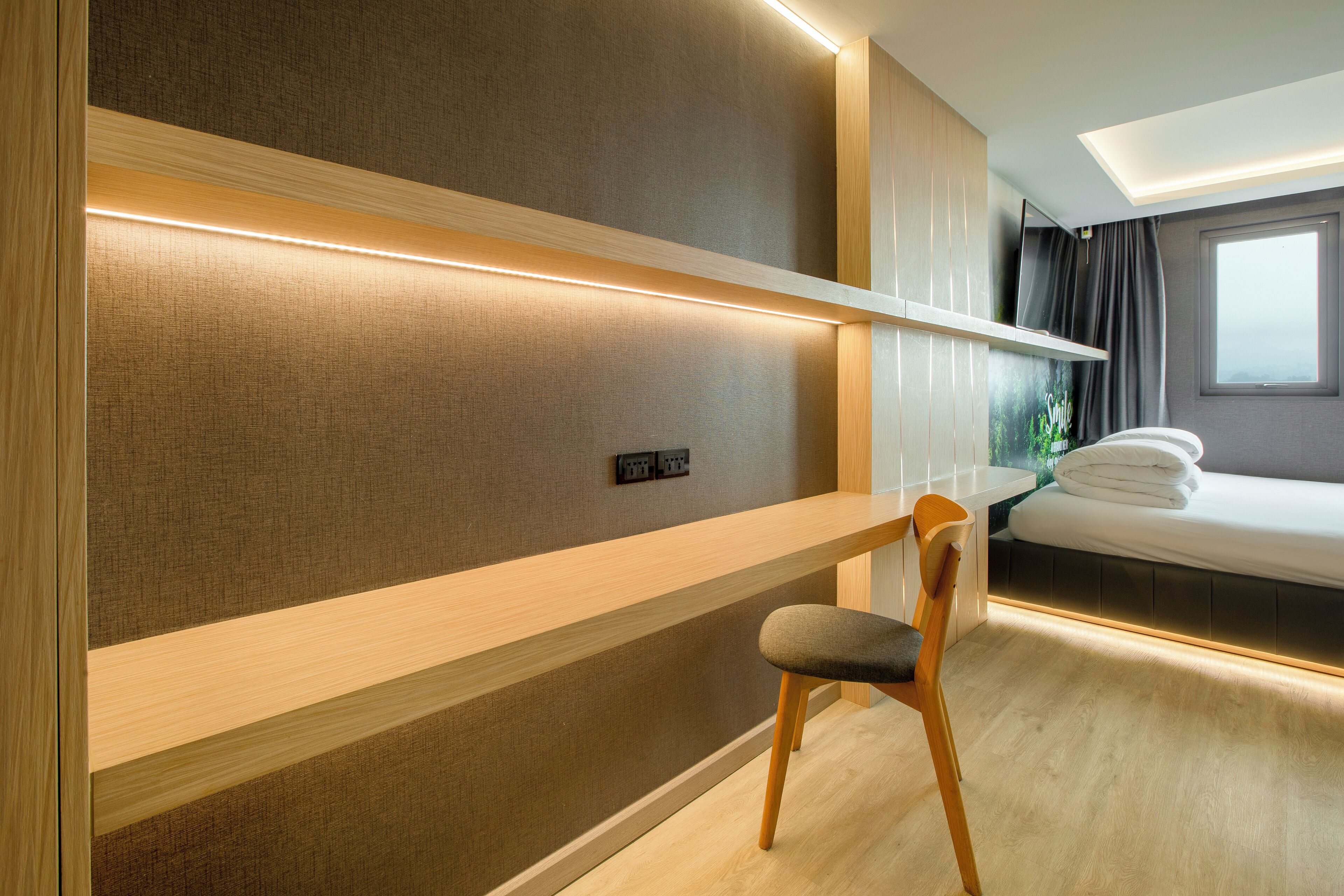 A modern hotel room with a wooden desk, a chair, a bed with a neatly made white duvet, and a window with curtains.