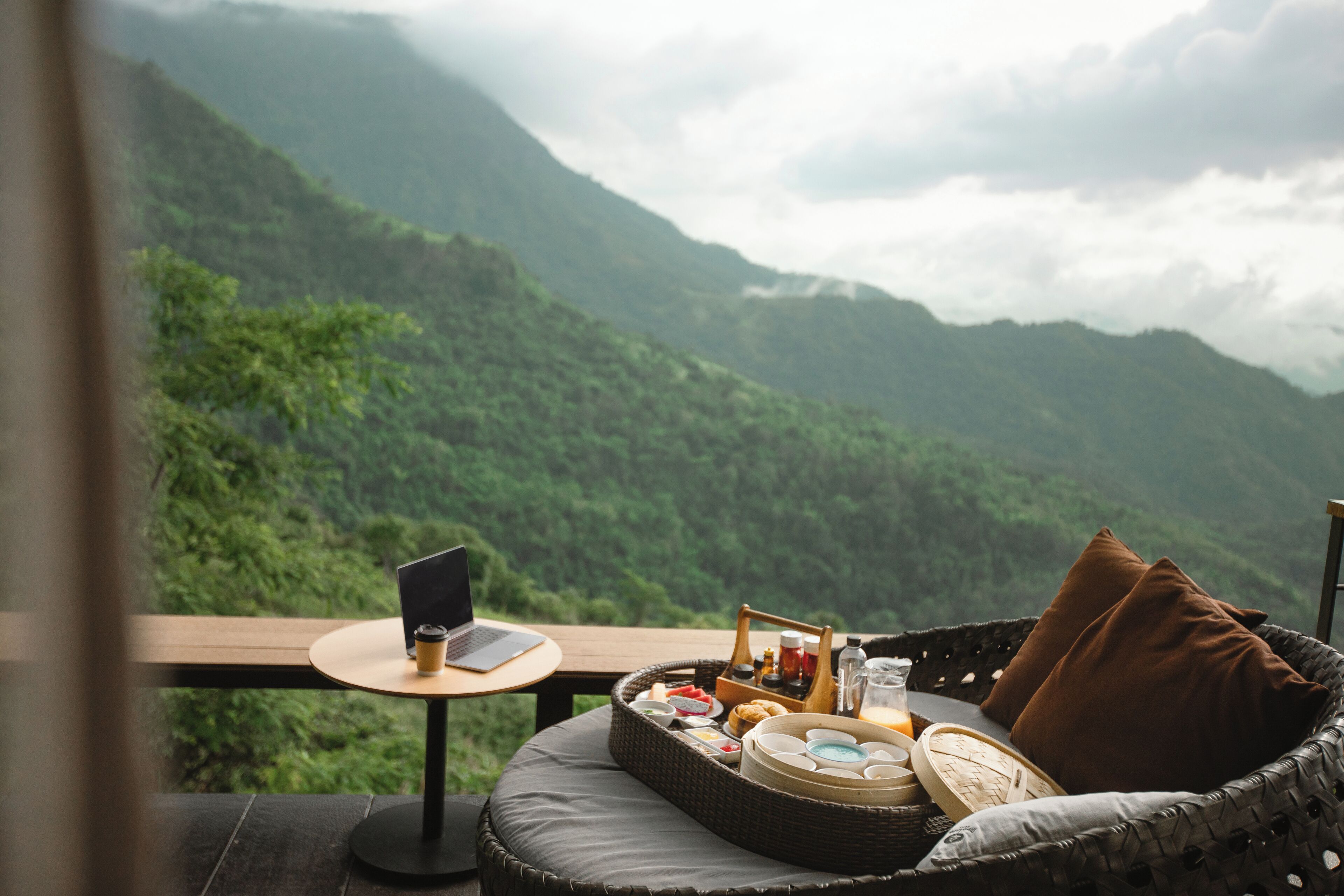 Una silla de mimbre con una computadora portátil y una cesta de alimentos en un balcón con vistas a un paisaje montañoso.