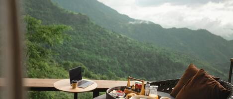 Una silla de mimbre con una computadora portátil y una cesta de alimentos en un balcón con vistas a un paisaje montañoso.