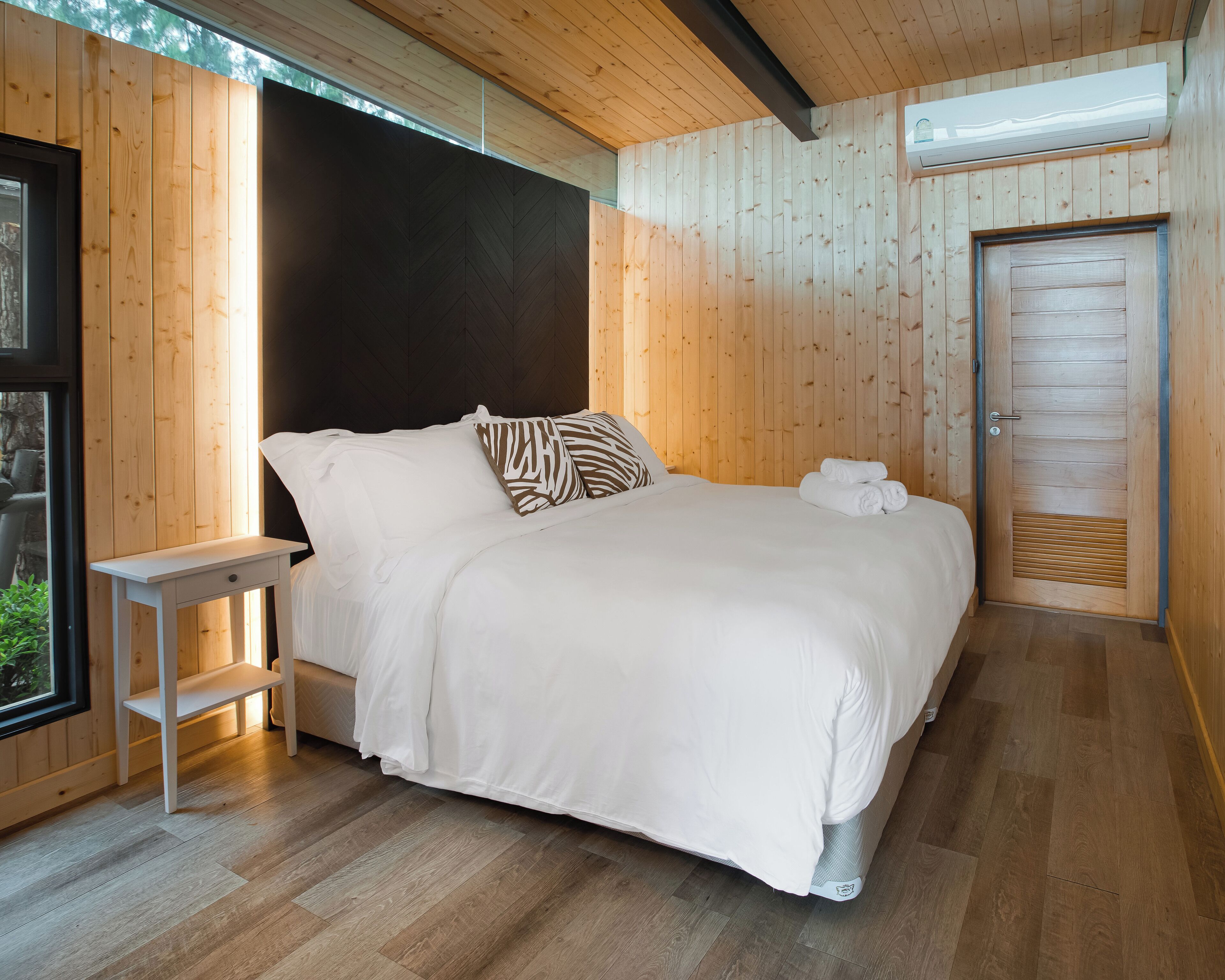 A bedroom with a bed, pillows, a bedside table, a window, and an air conditioning unit.